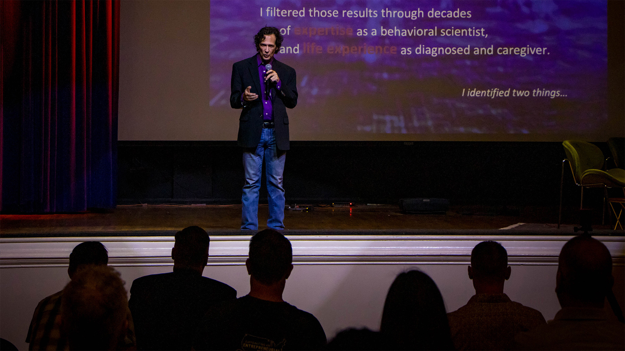 Dr. Payne on stage with a microphone, mid-sentence, speaking to a seated audience with a presentation slide lit behind him.