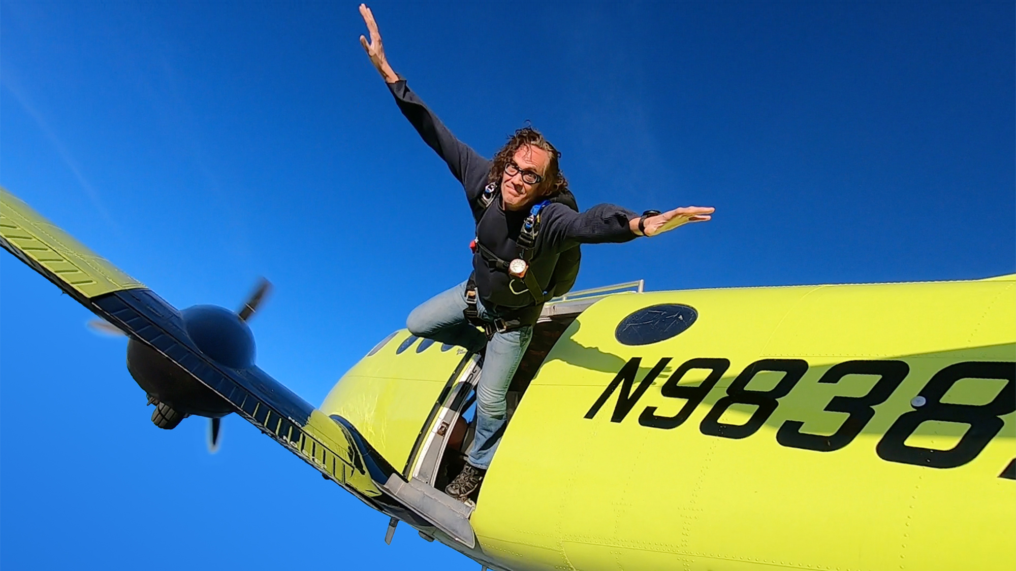 Dr. Kevin J. Payne standing in the open door of a yellow jump plane at altitude, arms out, looking toward the camera just before exit