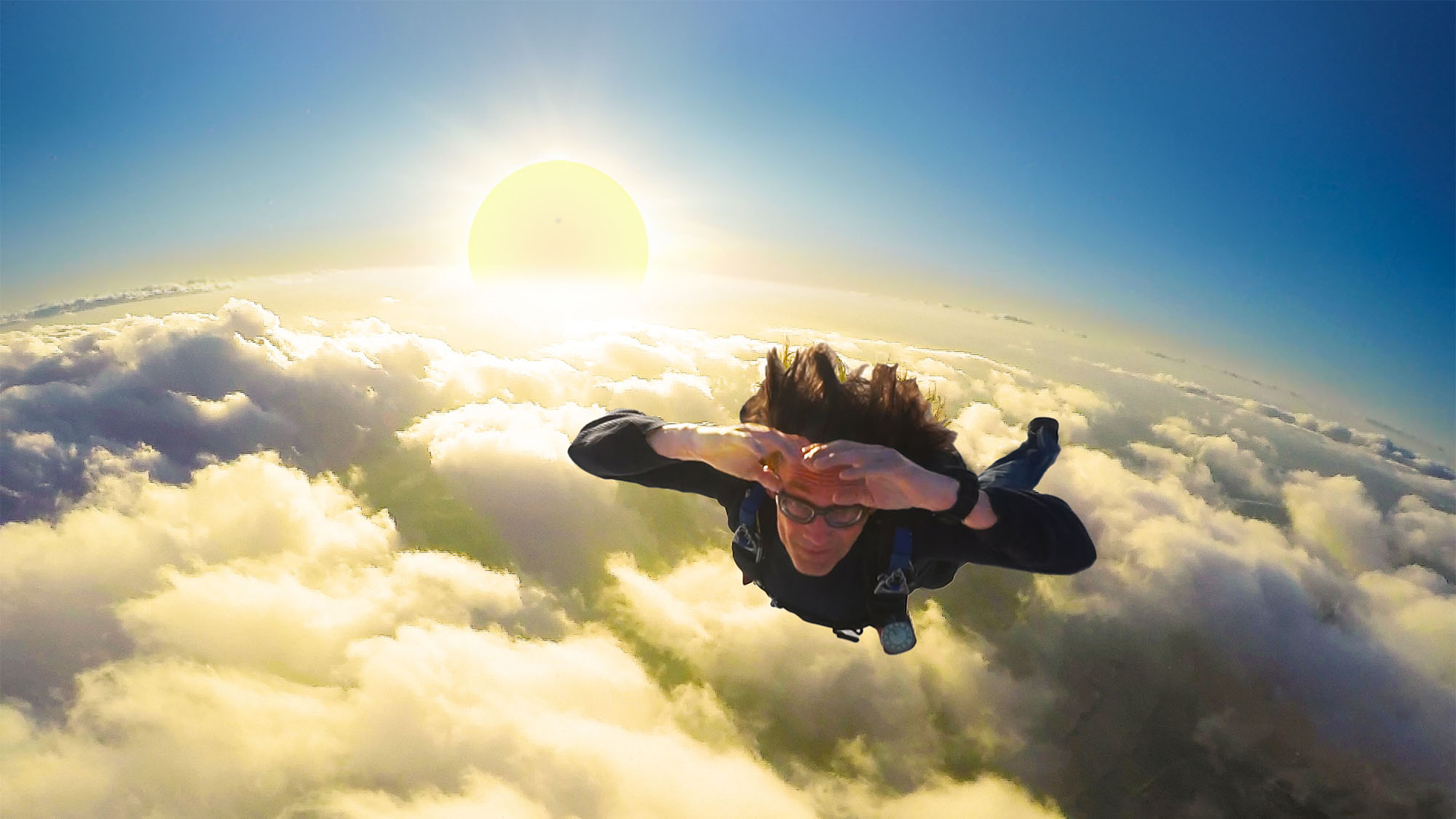 Dr. Kevin J. Payne in freefall above a sunlit cloud layer, hands at his forehead in a wave-off.